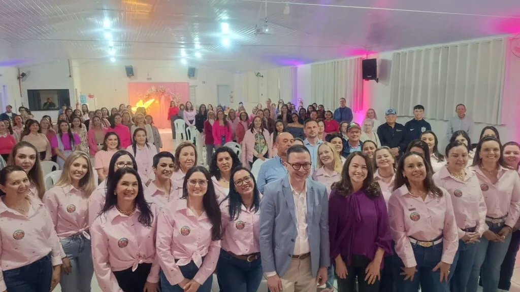 Foto coletiva dos participantes do encontro Outubro Rosa em Pinhão, em um salão com iluminação rosa. Na primeira fila, mulheres com camisas rosa posam ao lado de um homem de blazer azul e uma mulher de blusa roxa. Ao fundo, o público ocupa o espaço entre as cadeiras.