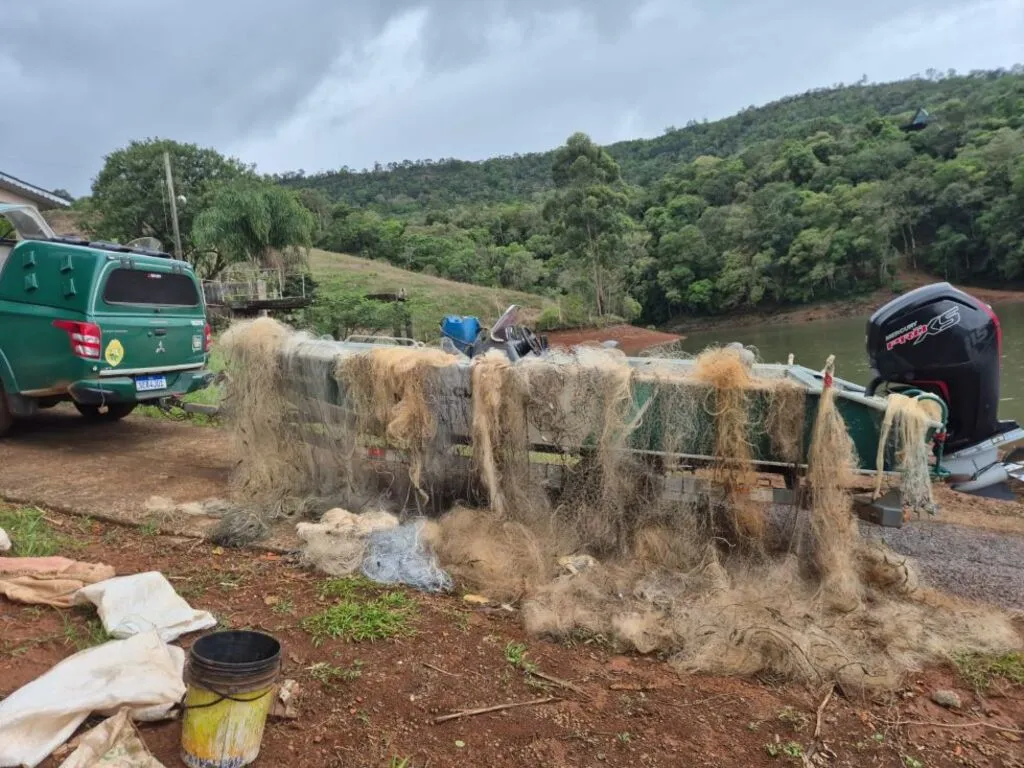 Redes de pesca apreendidas pendem das laterais de uma embarcação sobre carreta e se acumulam no chão, ao lado de uma viatura da Polícia Ambiental, às margens de uma represa.