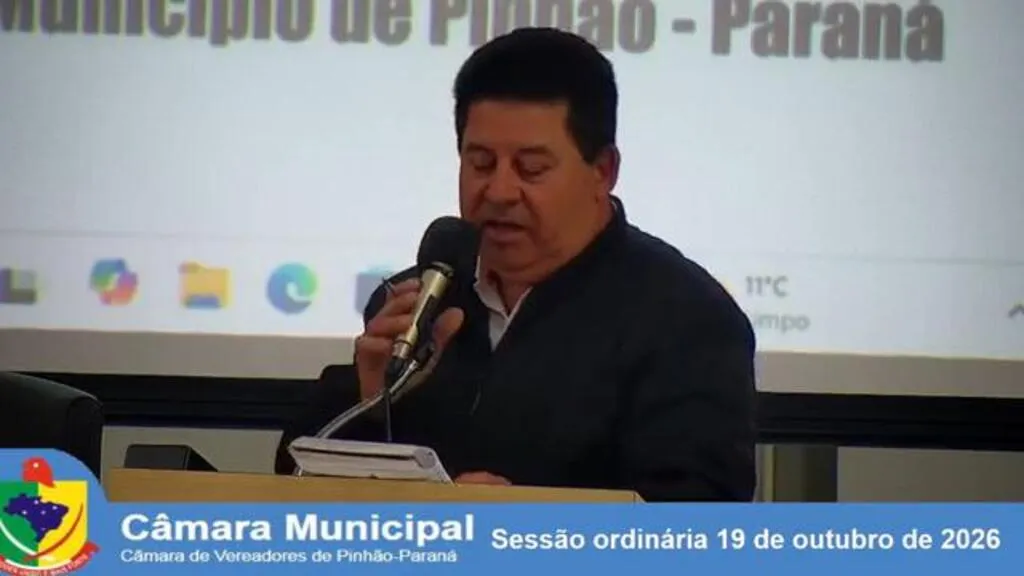Vereador Aroldo Antunes Domingues fala ao microfone na tribuna da Câmara Municipal de Pinhão, com documentos à sua frente. Na parte inferior, uma faixa identifica a sessão ordinária de 19 de outubro de 2026.