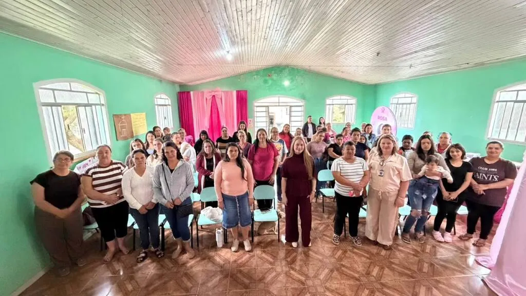 Mulheres reunidas para foto em um salão de paredes verdes, com cadeiras organizadas em fileiras e decoração rosa, durante encontro no bairro Dois Irmãos, em Pinhão.