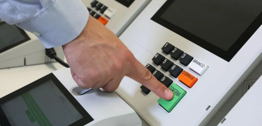 Mão de um eleitor pressionando a tecla verde “CONFIRMA” de uma urna eletrônica durante o processo de votação.