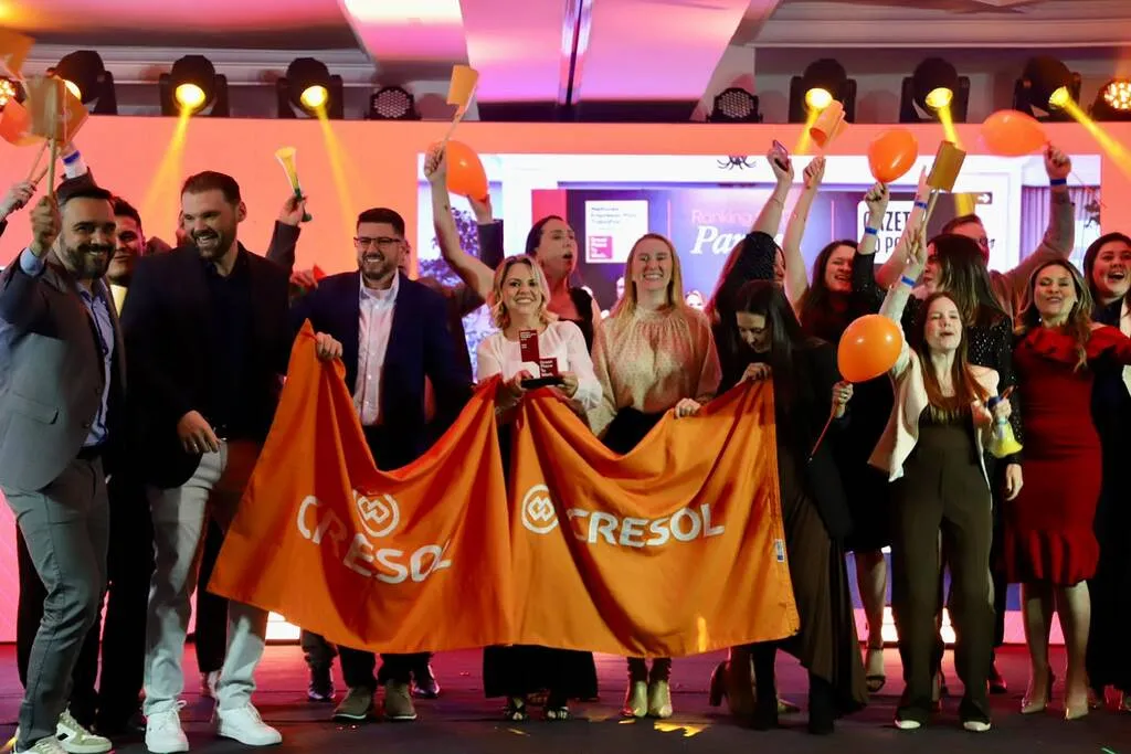 Grupo de pessoas comemora em um palco durante uma cerimônia de premiação. Participantes seguram bandeiras laranja da Cresol, balões e um troféu, sob luzes coloridas.