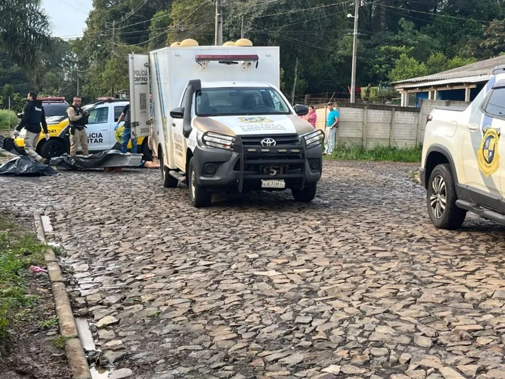 Viaturas da Polícia Científica e da Polícia Militar em uma rua de pedras no bairro Invernadinha, em Pinhão, com agentes próximos aos veículos e moradores ao fundo.