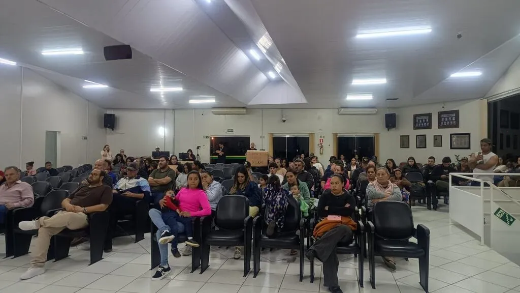 Moradores ocupam cadeiras no plenário da Câmara Municipal de Pinhão durante reunião sobre o abastecimento de água. Ao fundo, uma participante segura um cartaz com pedido de solução.