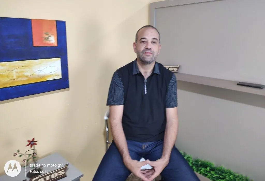 Maestro Rodrigo Correia Santos sentado no estúdio da TV Fatos durante entrevista sobre o Campeonato Paranaense de Fanfarras e Bandas.