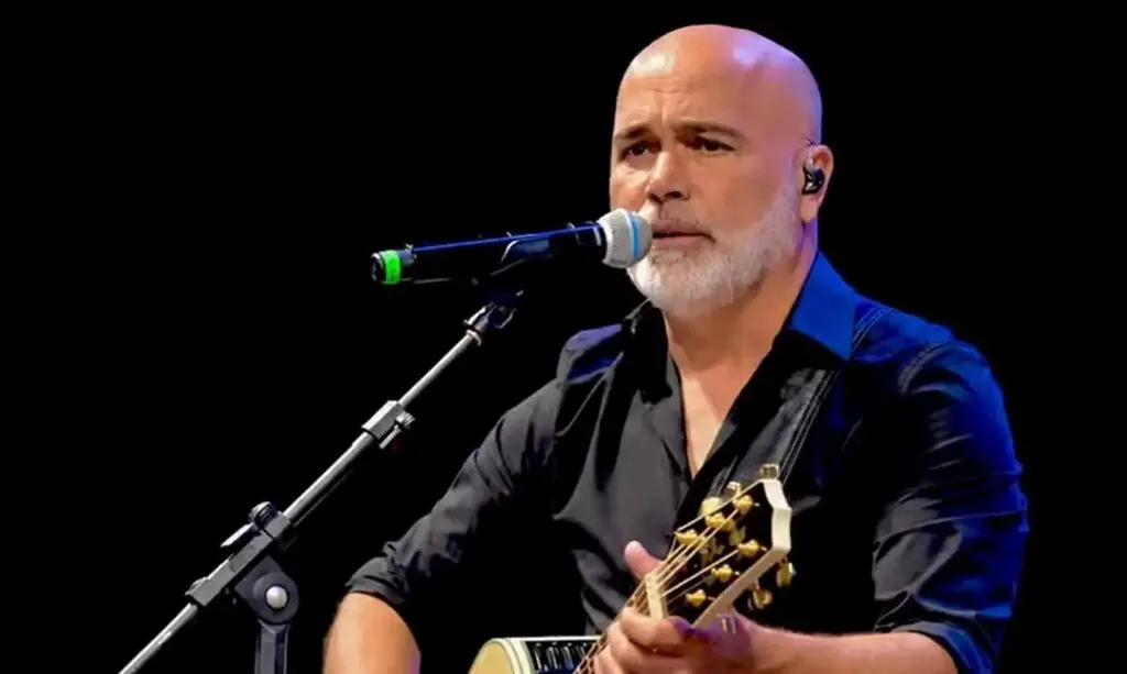 Cantor Rick se apresenta no palco, cantando ao microfone e tocando violão, durante um show.