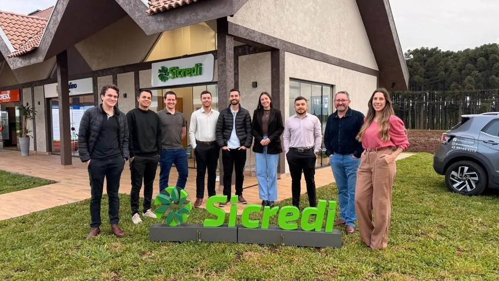 Nove pessoas, sete homens e duas mulheres, posam no gramado em frente a um espaço identificado com a marca Sicredi. À frente do grupo, há um letreiro verde com o nome e o símbolo da cooperativa. Ao fundo, aparecem um prédio com telhado de cerâmica e um veículo à direita.
