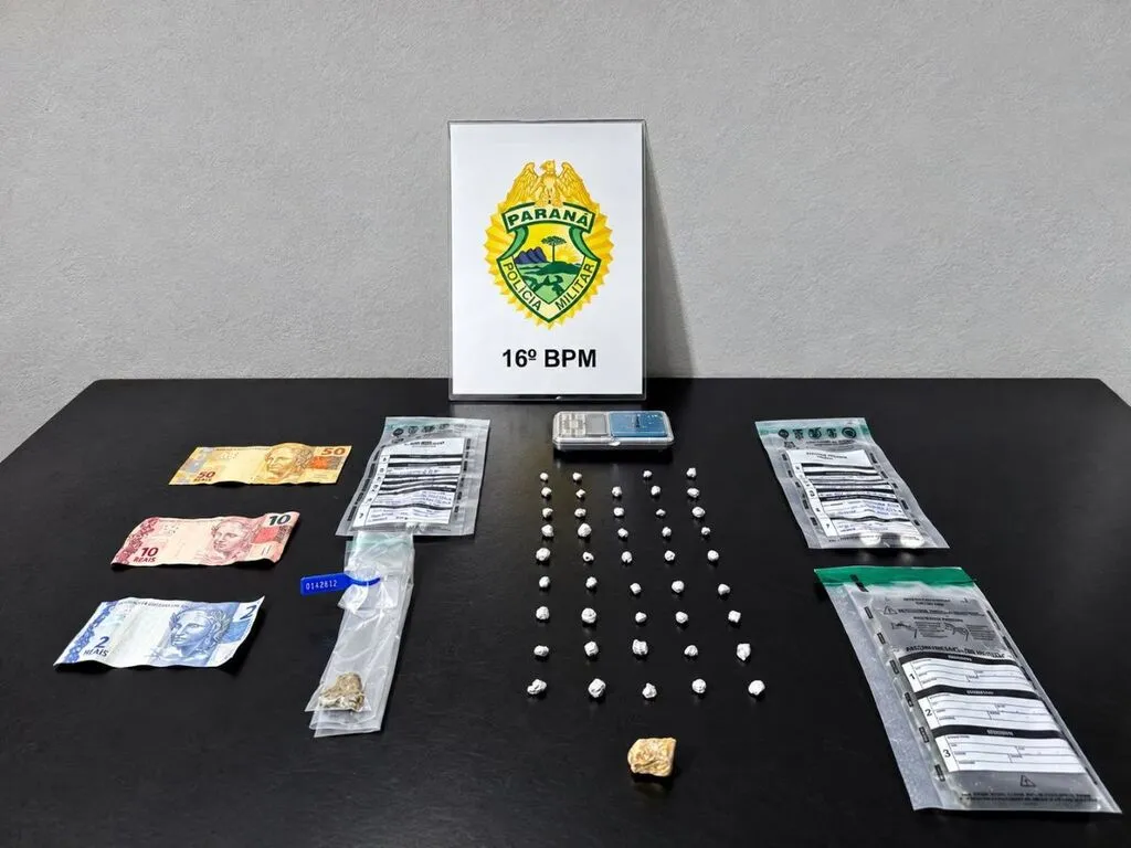 Materiais apreendidos pela Polícia Militar sobre uma mesa preta: pequenas porções embaladas, uma porção maior de substância amarelada, embalagens de evidências, balança de precisão e cédulas de R$ 50, R$ 10 e R$ 2. Ao fundo, placa com o brasão da PM do Paraná e a identificação do 16º BPM.