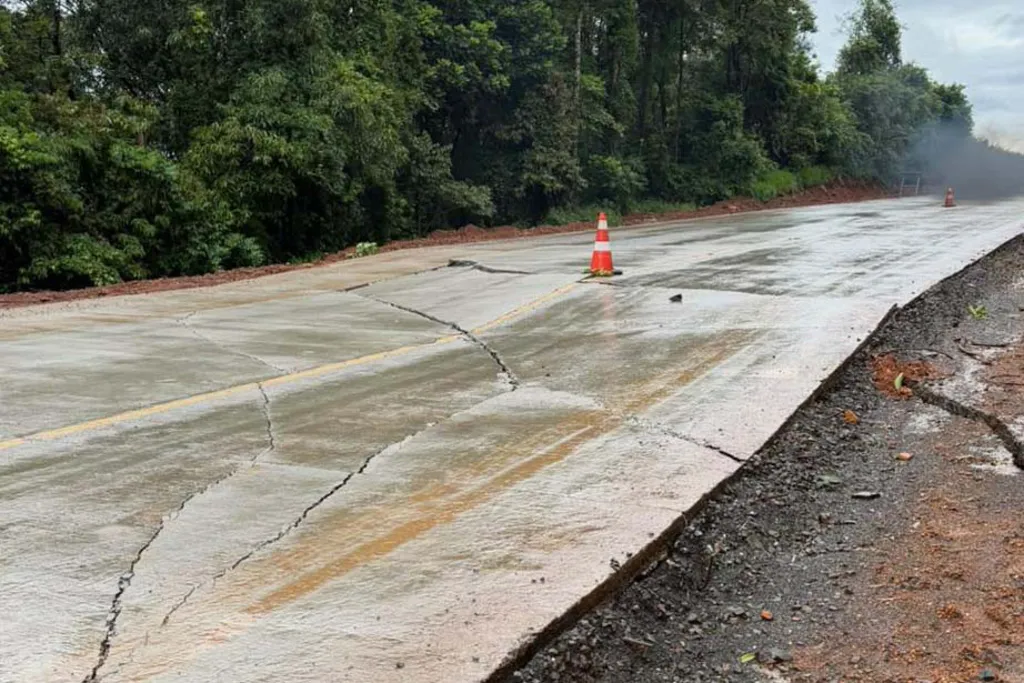 Foto ao nível da estrada mostrando uma rodovia pavimentada com rachaduras longitudinais e transversais extensas na pista. A superfície apresenta-se húmida, ladeada por vegetação densa e bermas de terra. Um cone de trânsito laranja está colocado sobre a faixa de rodagem danificada para alertar os condutores.