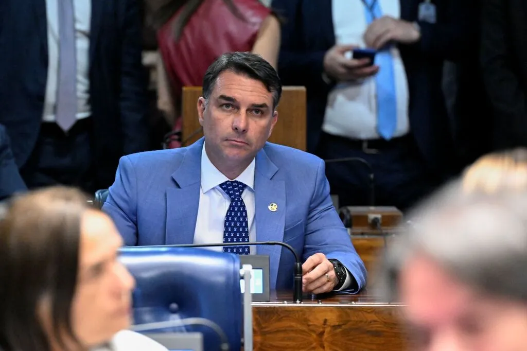 Flávio Bolsonaro sentado à bancada do plenário do Senado Federal, com a mão próxima ao rosto.