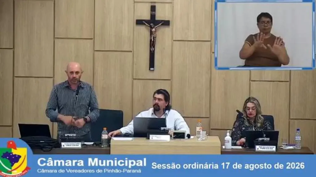 Vereador Luciano Padilha, em pé, defende a aprovação das contas de 2019 durante sessão da Câmara de Pinhão; à mesa estão João Paulo Mendes e Vilma Kitcky, com intérprete de Libras no telão.