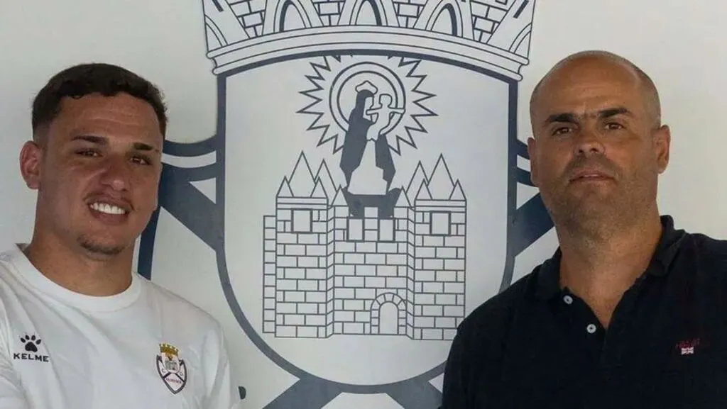 Jogador pinhãoense Adriel Moraes posa com a camisa do Feirense ao lado de um representante do clube, diante do escudo da equipe portuguesa.