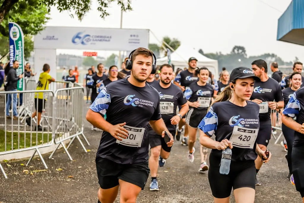 Fotografia em plano médio de um grupo de pessoas a correr numa rua durante um evento desportivo, com um pórtico de partida ao fundo com a inscrição "60 CORRIDAS SANEPAR". Em primeiro plano, destacam-se corredores com camisolas escuras com detalhes azuis e dorsais afixados (com os números 401, 420 e 128), sendo que o participante da esquerda usa auscultadores e o da direita segura numa garrafa de água.