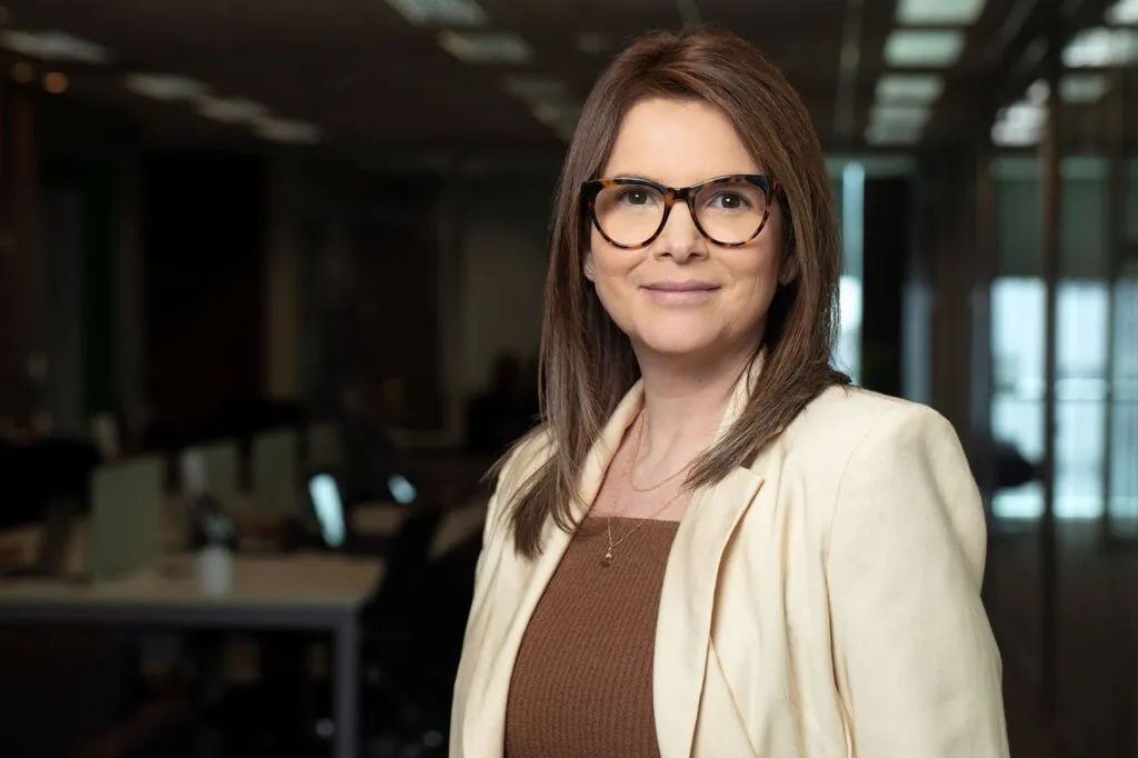 Mulher de cabelos castanhos e óculos posa em um escritório, usando blazer claro sobre uma blusa marrom. Ao fundo, mesas e computadores aparecem desfocados.
