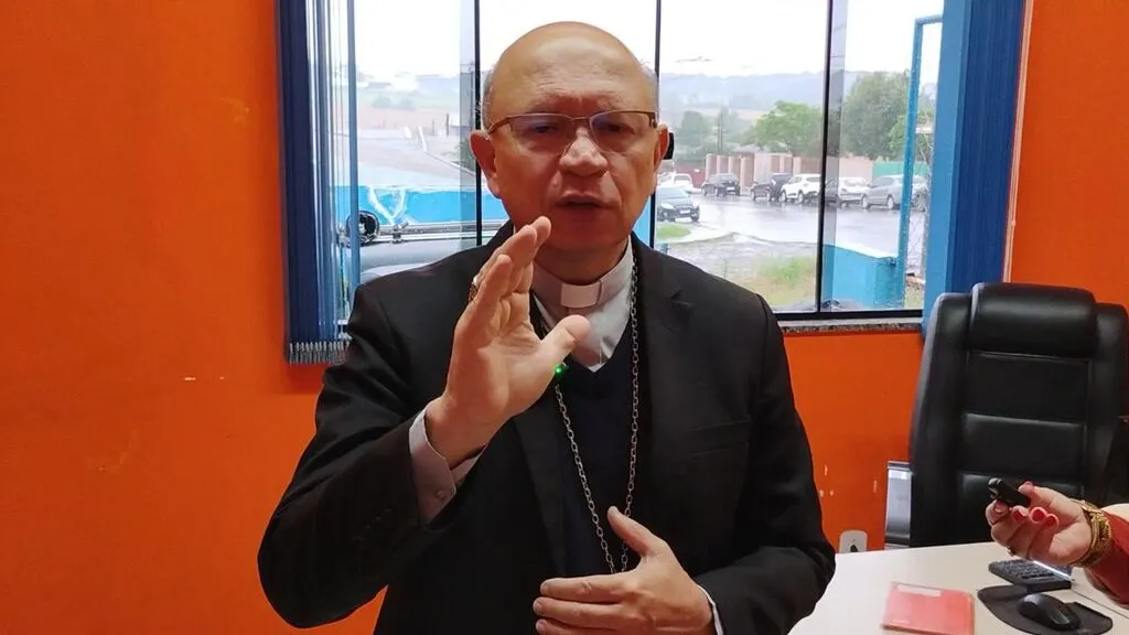 Dom Amilton Manoel da Silva, com vestes clericais e óculos, ergue a mão direita em gesto de bênção durante entrevista no posto da Brigada Comunitária de Pinhão.