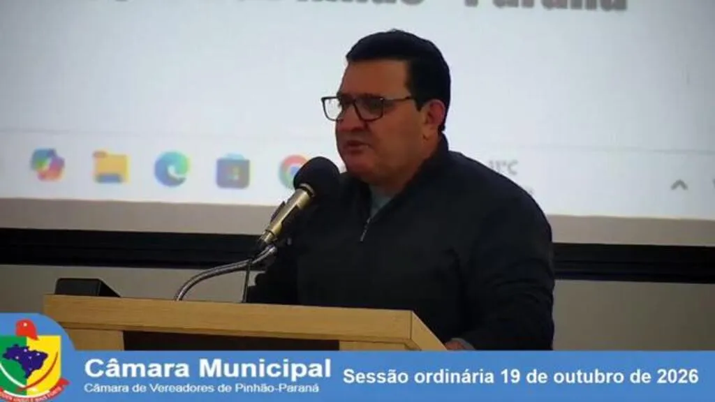 Vereador Alain Cesar de Abreu, de óculos e roupa escura, fala ao microfone na tribuna da Câmara Municipal de Pinhão. Na parte inferior, uma faixa identifica a sessão ordinária de 19 de outubro de 2026.