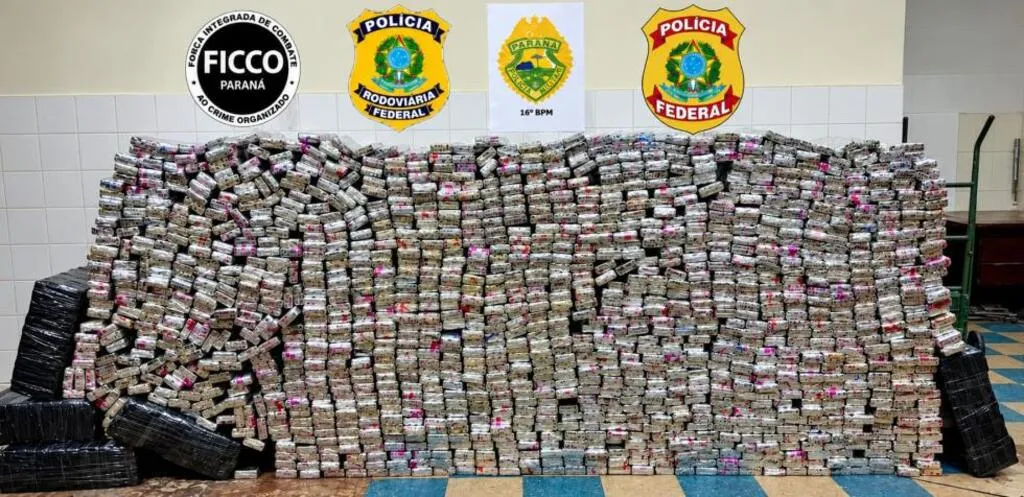 Centenas de tabletes de entorpecente empilhados sobre o chão, com volumes embalados em plástico preto nas laterais. Na parede, aparecem os símbolos da FICCO/PR, PRF, 16º BPM e Polícia Federal.