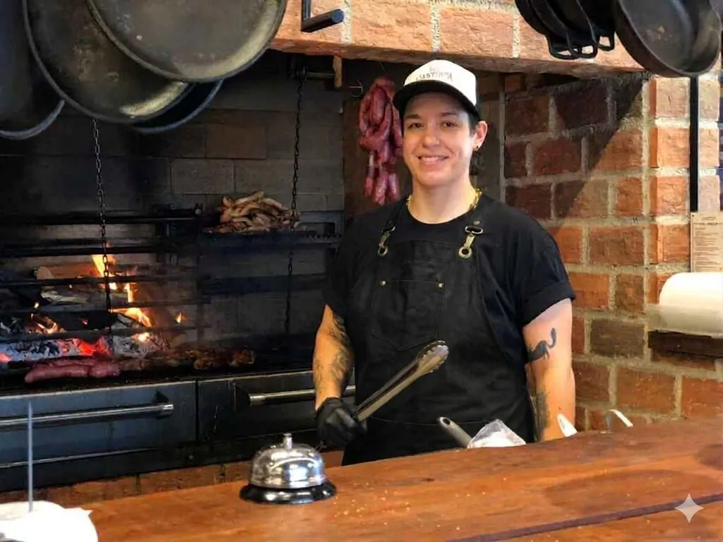 Churrasqueira Shirlei em frente a uma churrasqueira a fogo, segurando um pegador de carne, em ambiente rústico com panelas de ferro.