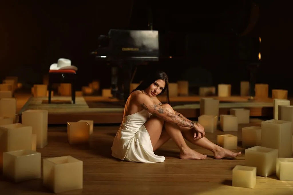 Ana Castela sentada no chão de madeira, usando vestido branco e descalça, cercada por luminárias de luz amarelada. Ao fundo, há um piano de cauda e um chapéu branco sobre uma banqueta.