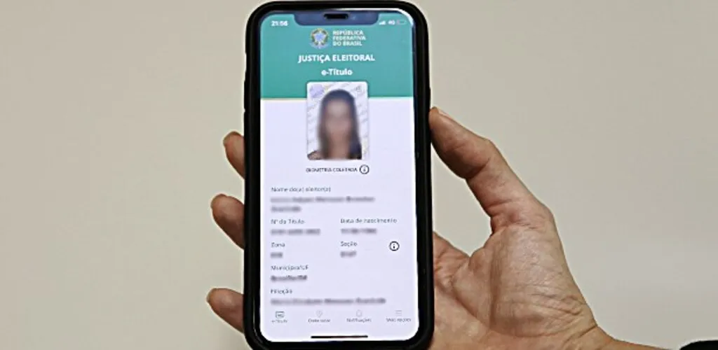 Mão segura um smartphone com o aplicativo e-Título aberto na tela, exibindo a versão digital do título de eleitor, com fotografia e dados cadastrais do eleitor.
