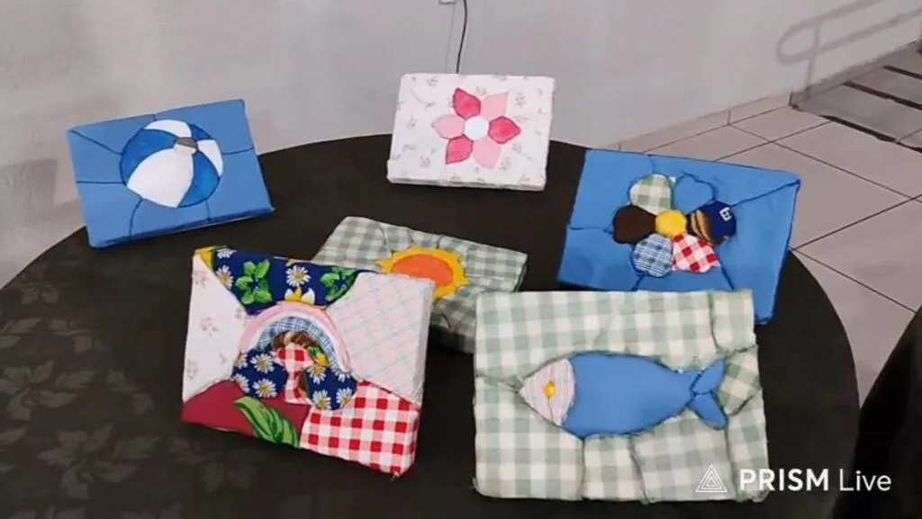 Quadros artesanais produzidos com retalhos de tecidos coloridos, representando flores, bolas, um peixe e outras figuras, expostos sobre uma mesa.