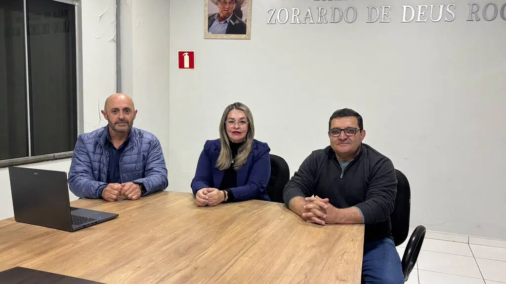 Três integrantes da CEI sentados à mesa na Câmara Municipal de Pinhão, com Vilma Kitcky ao centro, entre dois vereadores. Um notebook aberto está sobre a mesa, à esquerda.