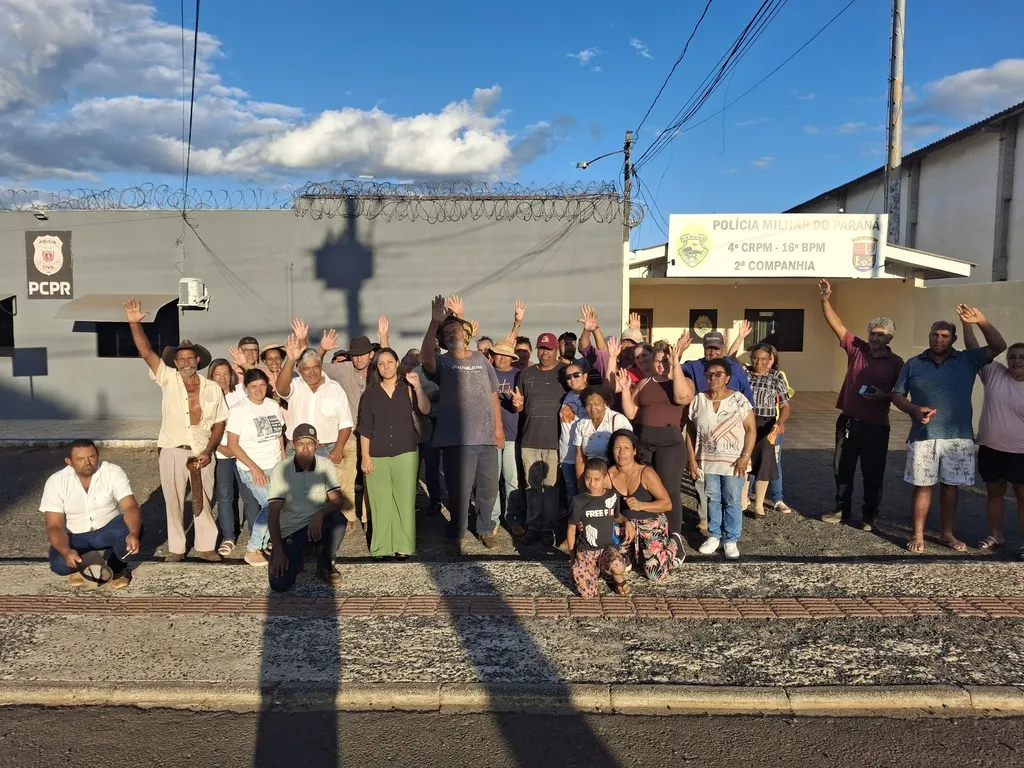 Integrantes da comunidade quilombola Paiol de Telha reunidos na calçada em frente às instalações da Polícia Civil e da Polícia Militar, em Pinhão. Várias pessoas estão com as mãos levantadas.