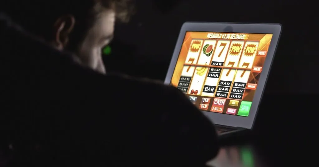 Pessoa vista de costas diante de um notebook que exibe um jogo de caça-níqueis on-line, com símbolos de frutas e números, em ambiente pouco iluminado.