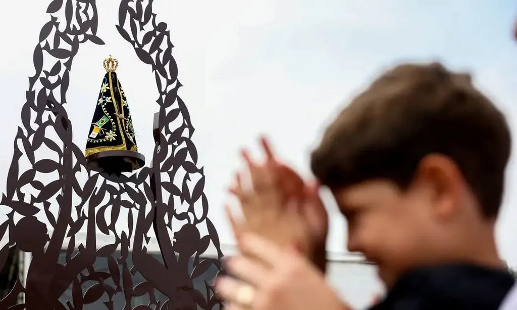 Imagem de Nossa Senhora Aparecida com coroa dourada e manto azul bordado, em uma estrutura metálica decorativa. À direita, uma pessoa desfocada aparece com a cabeça inclinada e as mãos unidas em oração.