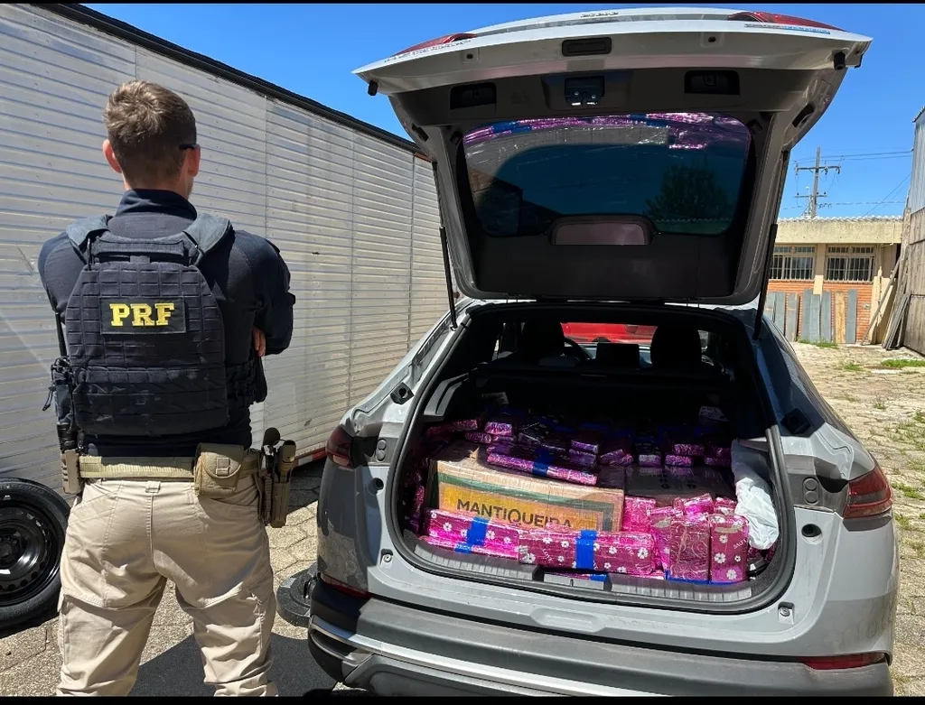 Foto em plano médio traseiro de um agente da Polícia Rodoviária Federal (PRF), com colete tático preto e a inscrição "PRF", a observar a bagageira aberta de um automóvel cinzento. O interior da mala está completamente preenchido com volumes embalados em papel de embrulho cor-de-rosa e caixas de cartão, destacando-se uma com a inscrição "MANTIQUEIRA".