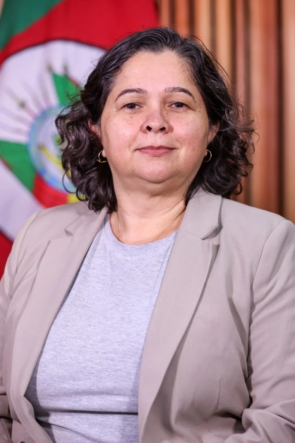 Pricilla Santana veste blazer bege sobre blusa cinza e posa diante da bandeira do Rio Grande do Sul e de um painel de madeira.