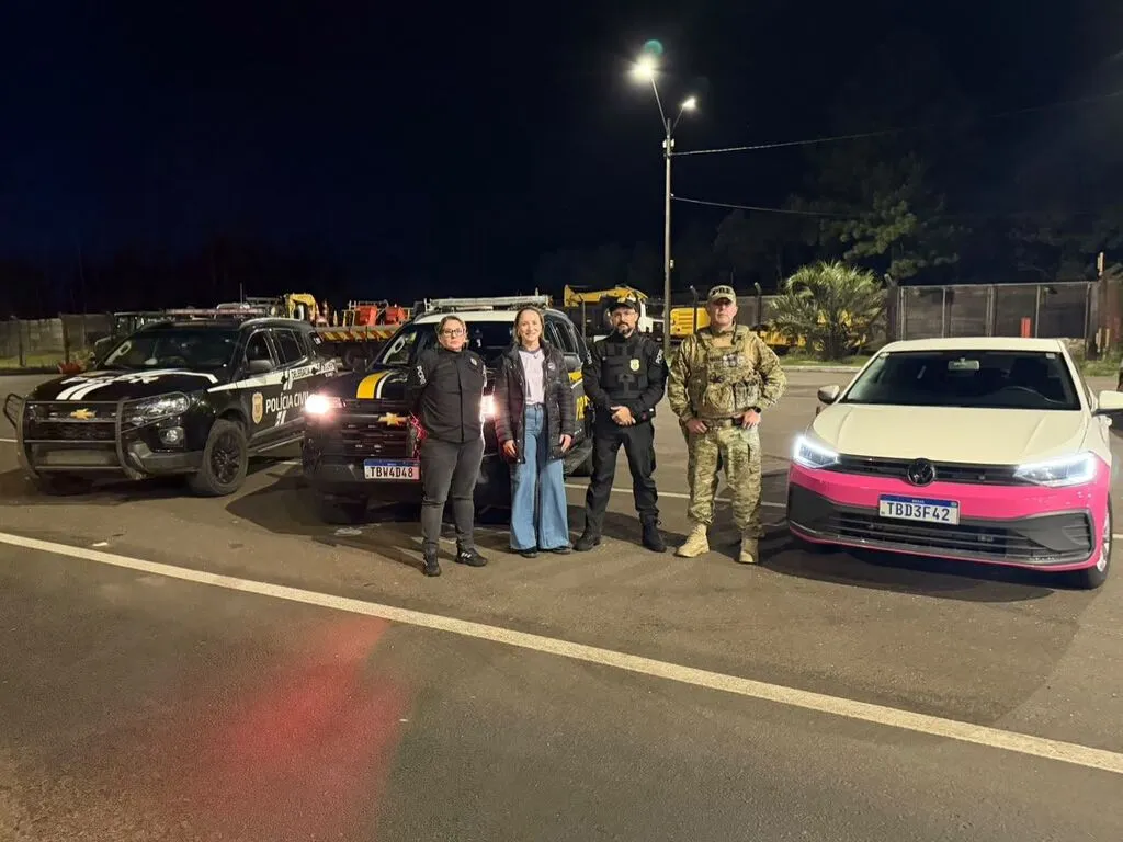 Quatro integrantes da operação Viagem Segura para Elas posam à noite entre viaturas da Polícia Civil, da PRF e um automóvel branco com faixa rosa, em Guarapuava.