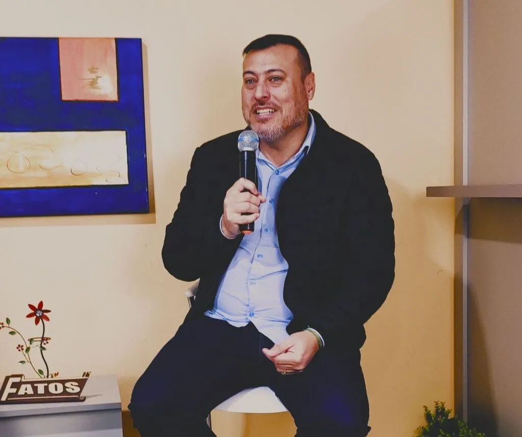Marcel, gerente da Cresol em Pinhão, sentado em uma banqueta no estúdio da TV Fatos, segurando um microfone. Ele veste camisa azul-clara e jaqueta preta, com um quadro e uma identificação do Fatos ao fundo.