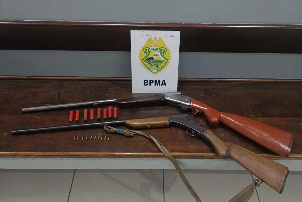 Duas armas de fogo de cano longo e munições dispostas sobre um banco de madeira, diante de uma placa com o brasão da Polícia Militar do Paraná e a sigla BPMA.
