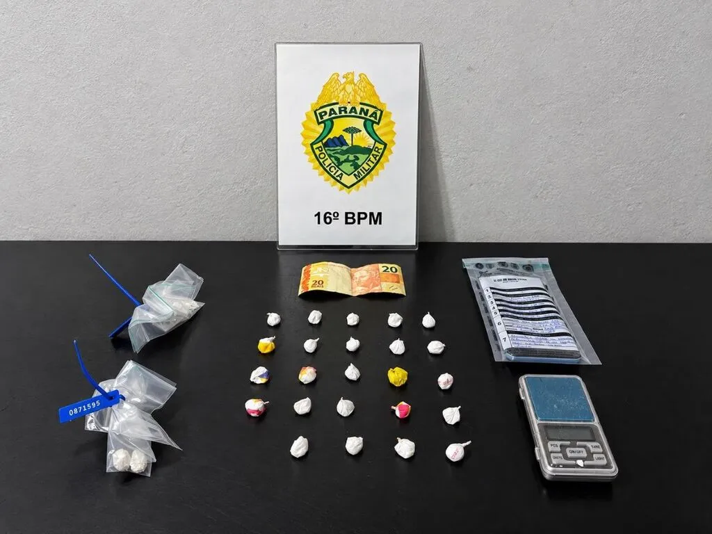 Invólucros de substância semelhante à cocaína, dinheiro, celular em embalagem lacrada e balança de precisão sobre uma mesa, diante de uma placa do 16º Batalhão da Polícia Militar do Paraná.