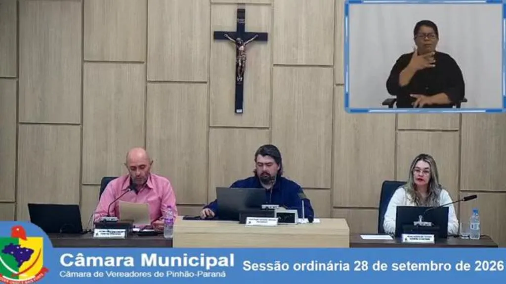 Mesa Diretora da Câmara Municipal de Pinhão durante a sessão ordinária de 28 de setembro de 2026. Três integrantes estão sentados à frente do plenário, e a transmissão exibe uma intérprete de Libras no canto superior direito.