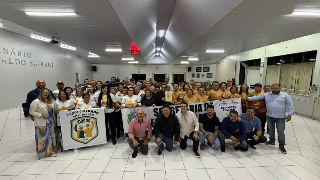 Grupo de atletas idosos, profissionais e vereadores posa no plenário da Câmara Municipal de Pinhão após a entrega da Moção de Congratulações. Os participantes seguram faixas da equipe, e uma pessoa ao centro exibe a homenagem