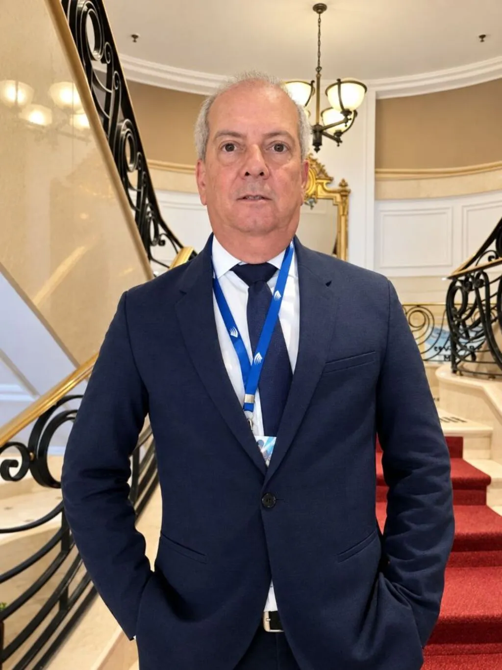 Marcos da Silva Ramos, de terno azul-marinho, camisa branca e gravata, posa diante de uma escadaria com corrimãos ornamentados e carpete vermelho.