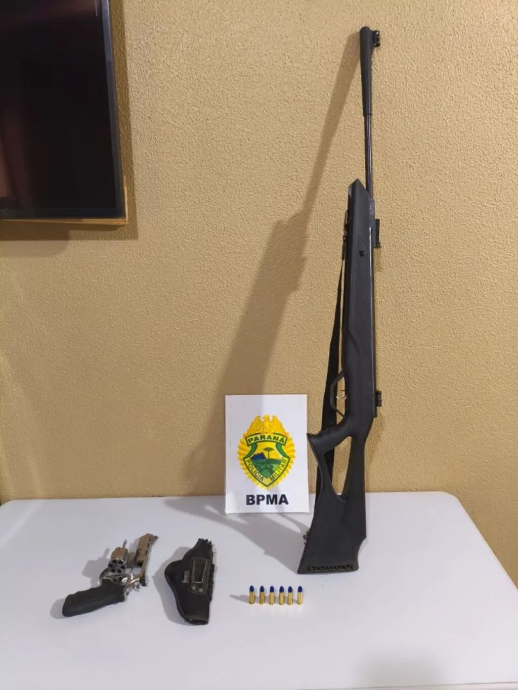 Espingarda de pressão, revólver, coldre e seis munições dispostos sobre uma mesa ao lado da identificação do Batalhão de Polícia Militar Ambiental.