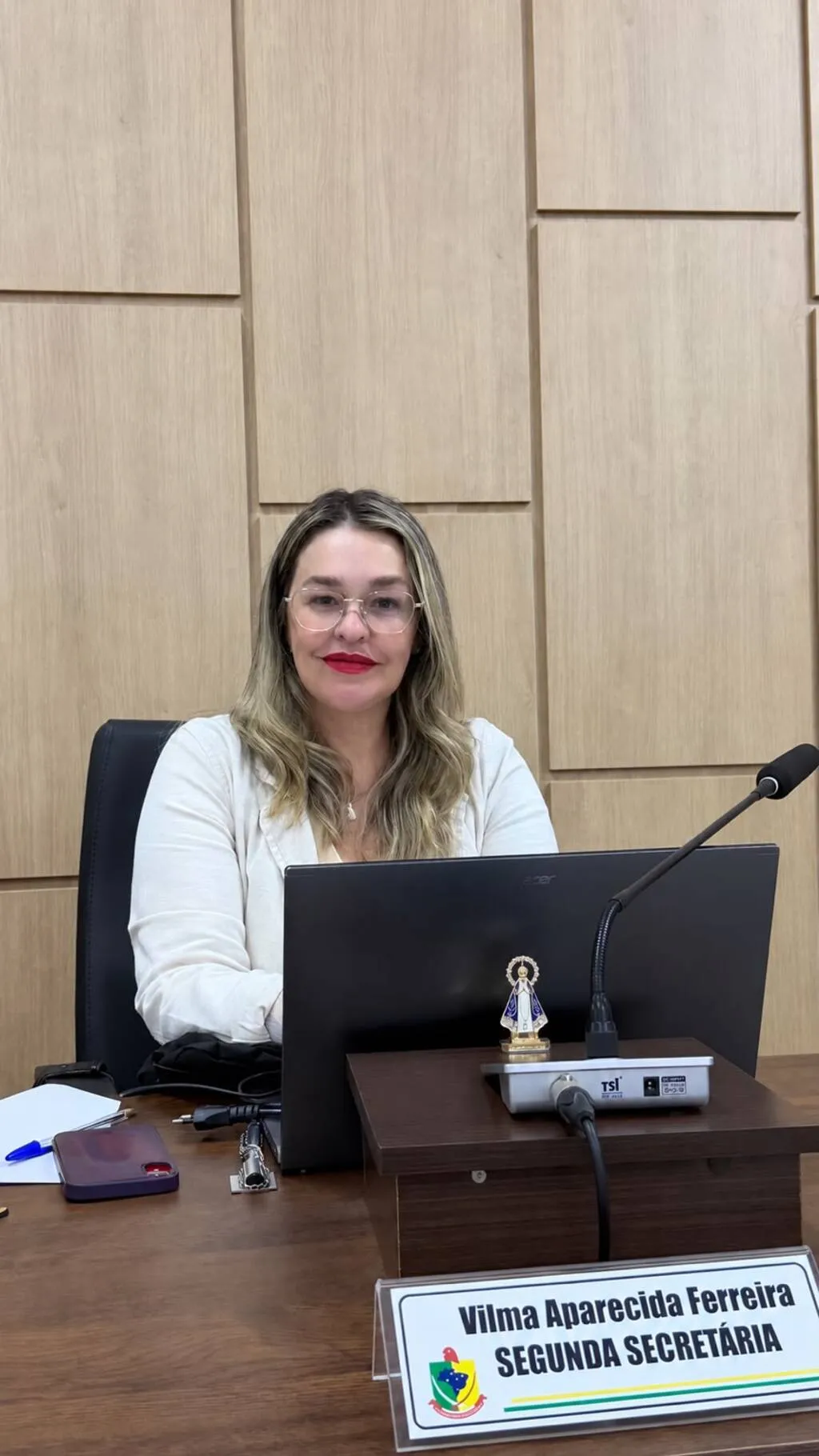 Vereadora Vilma Aparecida Ferreira, de óculos e blusa branca, sentada à mesa da Câmara Municipal de Pinhão, diante de um notebook e um microfone. À frente, uma placa a identifica como segunda secretária.