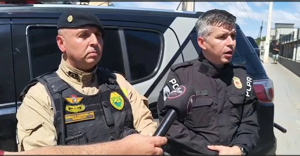 Representantes das polícias Militar e Civil concedem entrevista diante de uma viatura. À esquerda, um policial militar usa boina e colete; à direita, um policial civil fala ao microfone.