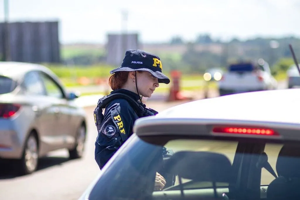 Policial rodoviária federal uniformizada ao lado de um automóvel, durante fiscalização, com outros veículos e cones ao fundo.