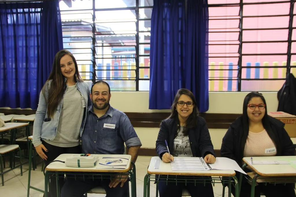 Quatro mesários posam para foto em uma seção eleitoral de Pinhão (PR), durante as eleições de 2018, sentados e em pé diante das mesas utilizadas para os trabalhos da votação.