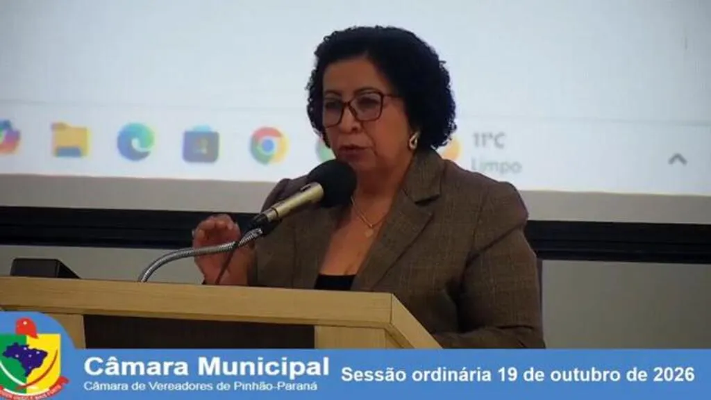 Vereadora Solange Adronski, de óculos e blazer marrom, fala ao microfone na tribuna da Câmara Municipal de Pinhão. Uma faixa na parte inferior identifica a sessão ordinária de 19 de outubro de 2026.
