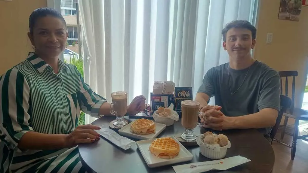 Foto em plano médio de dois adultos sentados à mesa em uma cafeteria. À esquerda, a jornalista Ana Ligia Sena usa uma camisa de listras verticais verdes e brancas. À direita, Francesco Buss Danieli, um dos proprietários da Cheirin Bão, veste camiseta cinza. Sobre a mesa escura redonda há dois combos compostos por xícaras de café com leite, pratos com waffles recheados e porções de pão de queijo.