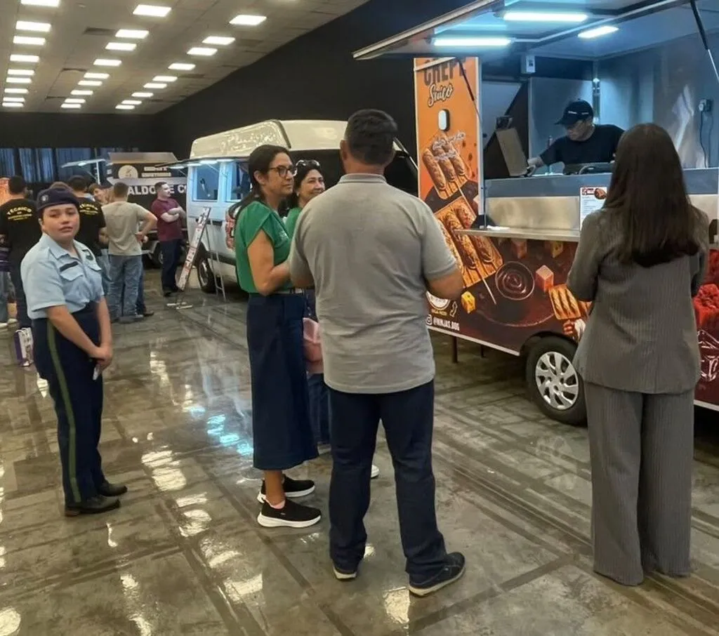 Foto em ambiente interno com piso brilhante, mostrando pessoas reunidas em frente a um food truck escuro adesivado com imagens de alimentos. À esquerda, uma jovem em pé vestindo uniforme escuro com boina e calça social observa o movimento. No centro, três pessoas conversam de costas para a câmera, enquanto à direita uma mulher de terno cinza observa o atendimento no balcão do veículo.