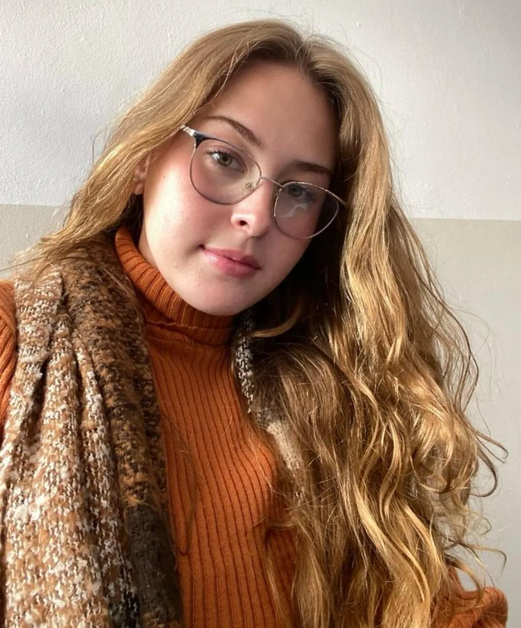Mariana Huller, colunista do Portal Fatos do Iguaçu, com cabelos longos e ondulados, usando óculos, blusa de gola alta em tom terracota e cachecol marrom, diante de uma parede clara.