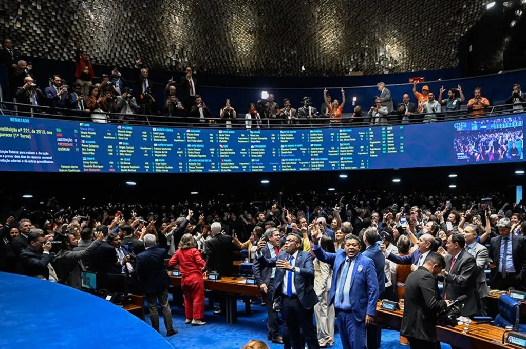 Plenário do Senado Federal lotado, com parlamentares de pé, alguns com os braços erguidos, diante do painel eletrônico de votação. Nas galerias, pessoas acompanham a sessão e registram o momento.
