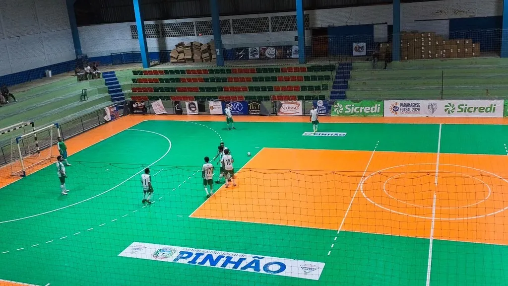 Jogadores de futsal em quadra verde e laranja, próximos à área de um dos gols, em ginásio com arquibancadas ao fundo e rede de proteção em primeiro plano.