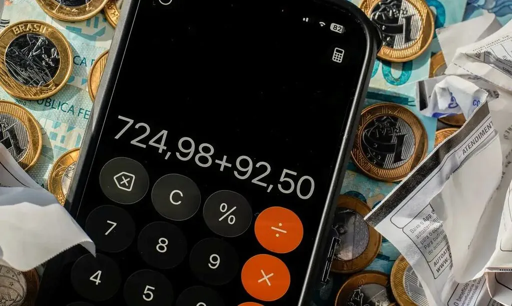 Celular com calculadora exibindo a soma “724,98 + 92,50”, sobre cédulas de reais, moedas de um real e comprovantes de papel amassados.